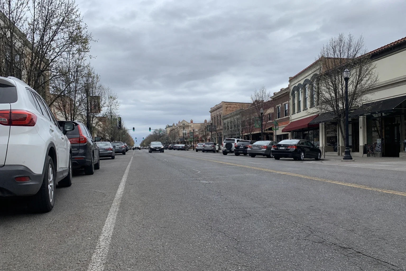 Low view down a downtown main street on an overcast day, with cars parked along both sides, bare trees, street lamps and two-story brick storefronts with awnings